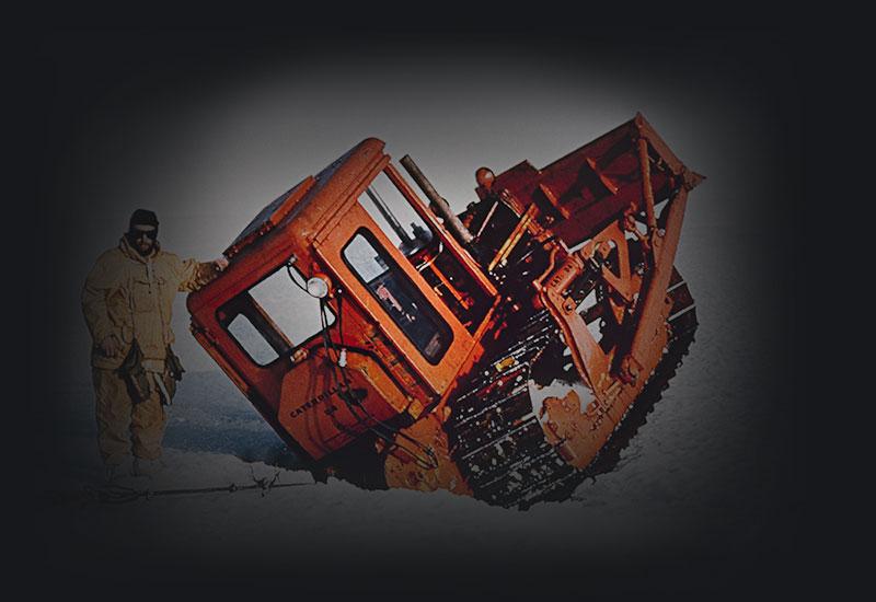 Man in yellow leaning against orange grader wedged in ice crevice.