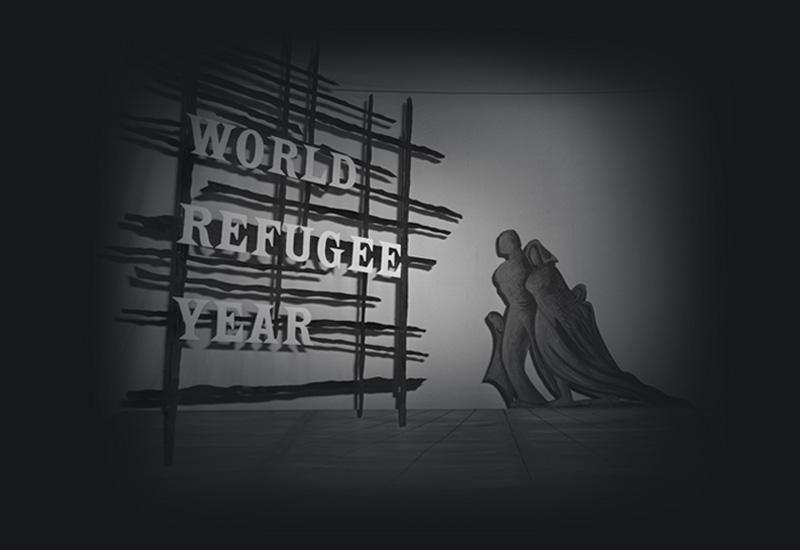 Sign reading 'World Refugee Year' next to an abstract statue of a family walking towards it.