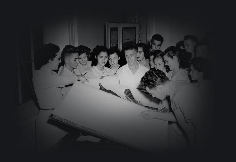A group of students gathered around a large book on a lectern. 