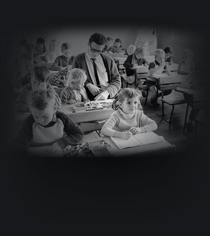 Young children sitting at desks in a classroom while a teacher assists one of the students.