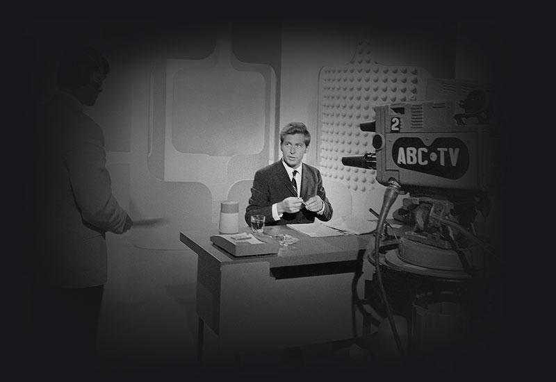 A man sitting at a desk infront of an ABC-TV camera.