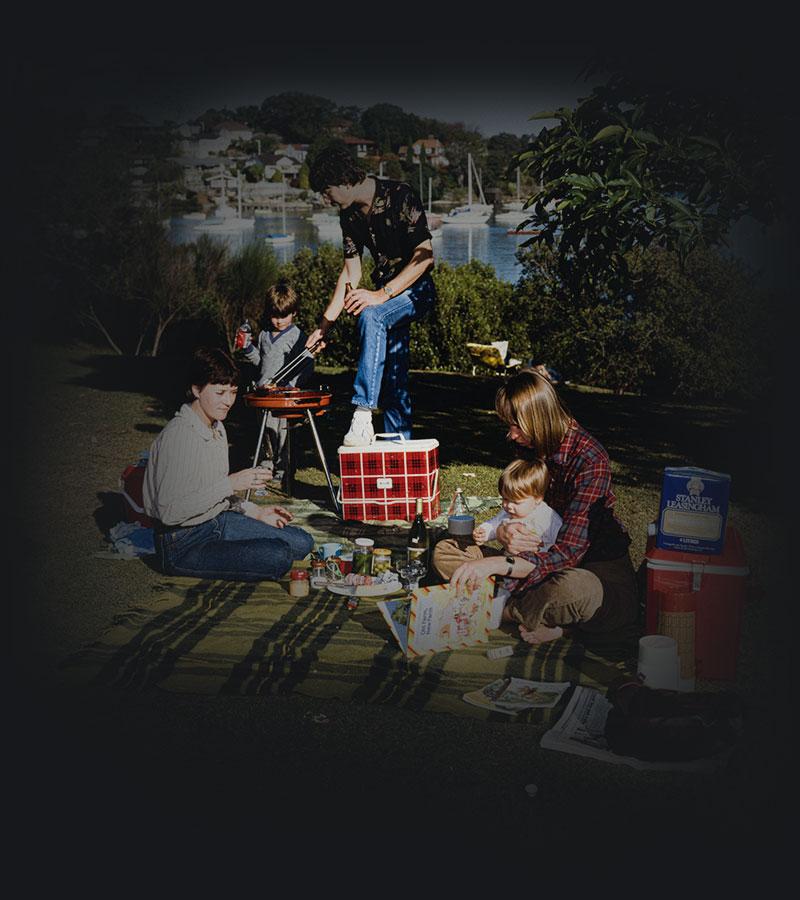 3 people and 2 small chidlren with a BBQ and picnic blanket on lawn with boats in the background.