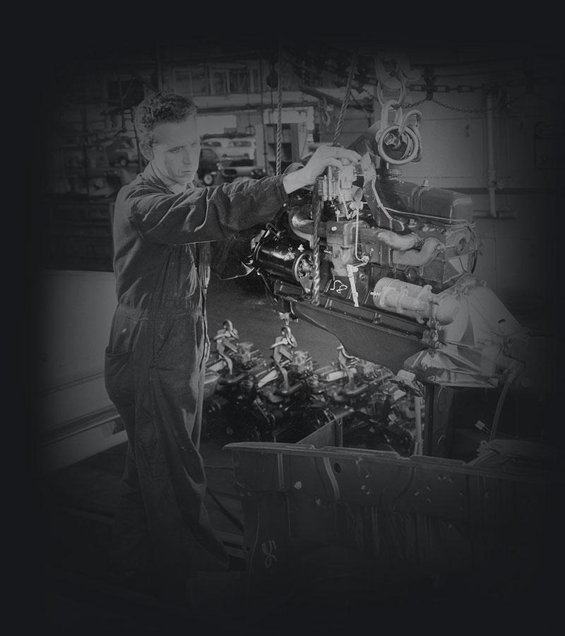 A production line worker guides an engine into the engine bay of a car body.