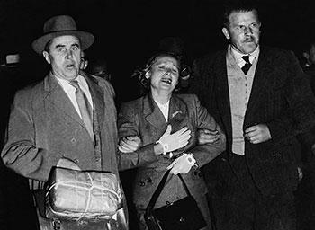 Black and white photograph: two men in coats escorting a woman by the arms.