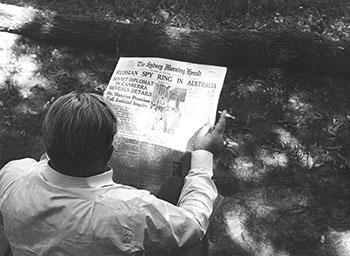 Black and white photograph: a man seated outside holding a newspaper and cigarette.