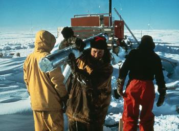 Four men in snow dressed in waterproof clothing. One carrying a silver foil wrapped ice core.