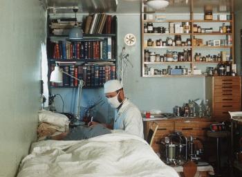 A doctor wearing white coat, cap and face mask seated beside a patient. Small room with shelving medicine and medical equipment.