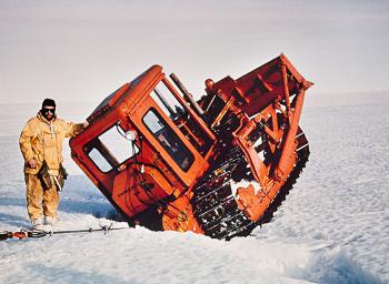 Man in yellow leaning against orange grader wedged in ice crevice.