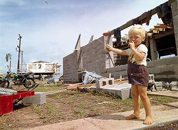 A boy with a red kite, a green trail bike and 2 destroyed buildings.