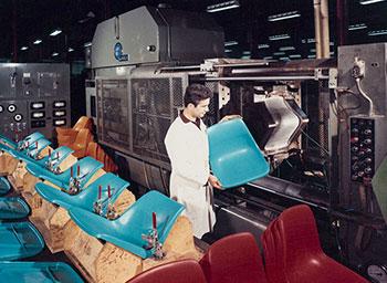 A man holds a blue plastic chair seat beside a large injection moulding machine