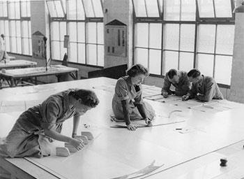 2 women sit on a table to make large workshop drawings.