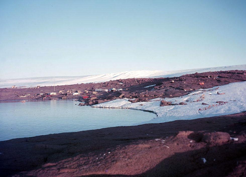Mawson station in 1955. Buildings next to the sea, surrounded by snow and rock.