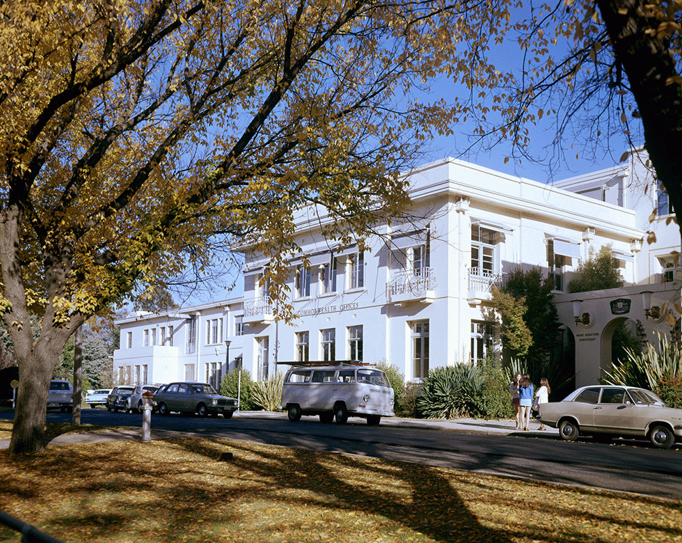 A view from beneath trees in autumn of people standing beside East Block building.
