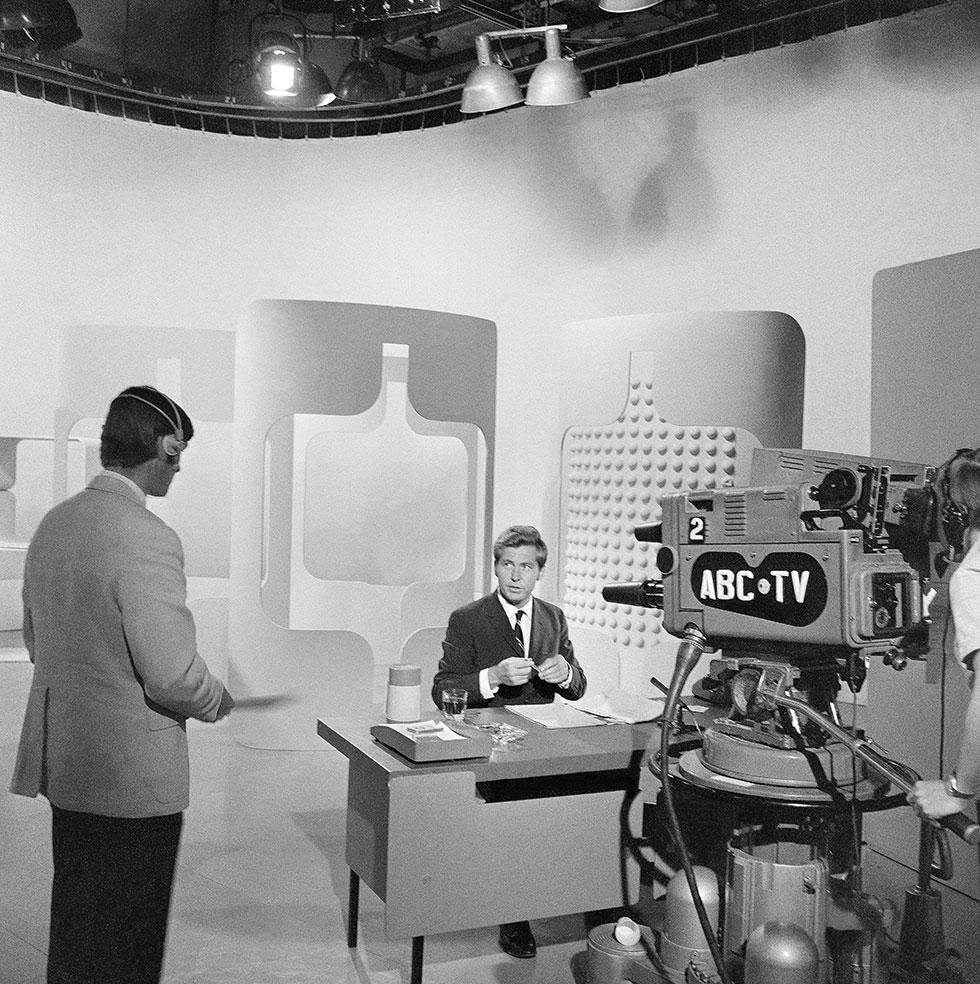 A man sitting at a desk infront of an ABC-TV camera.