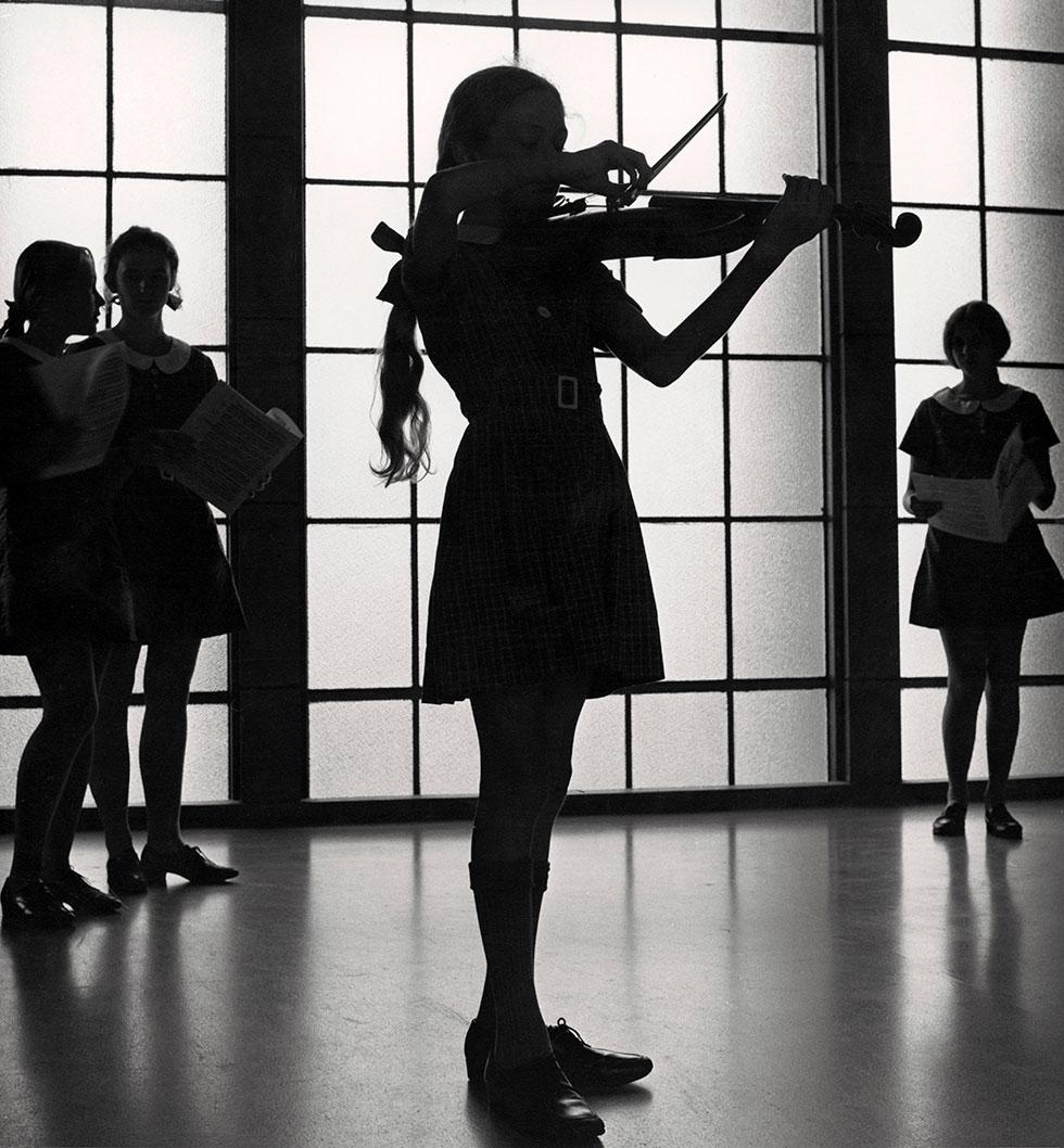 Girl playing violin while silhouetted infront of a window while others stand and watch