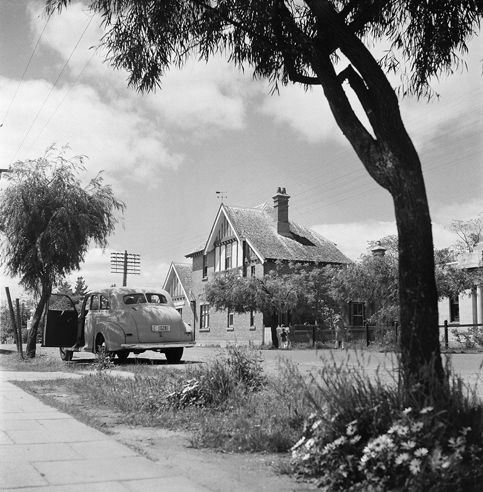 1950s car parked with open door beside a tree near a 2 story building.