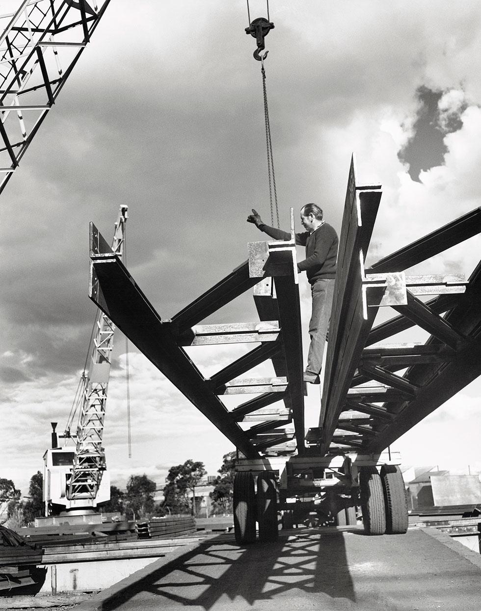 A man stands on steel trusses being lifted by a crane.