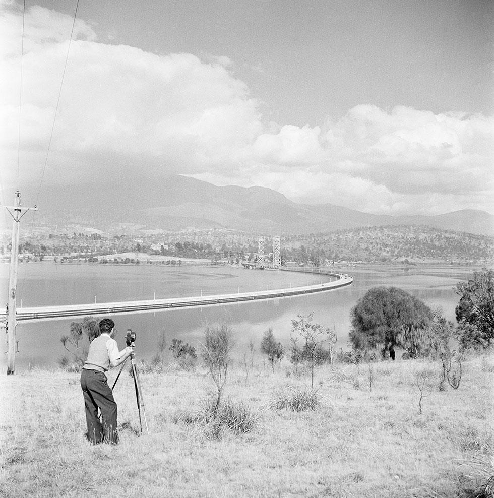 A man with a motion film camera and tripod looks down across a river and pontoon bridge.