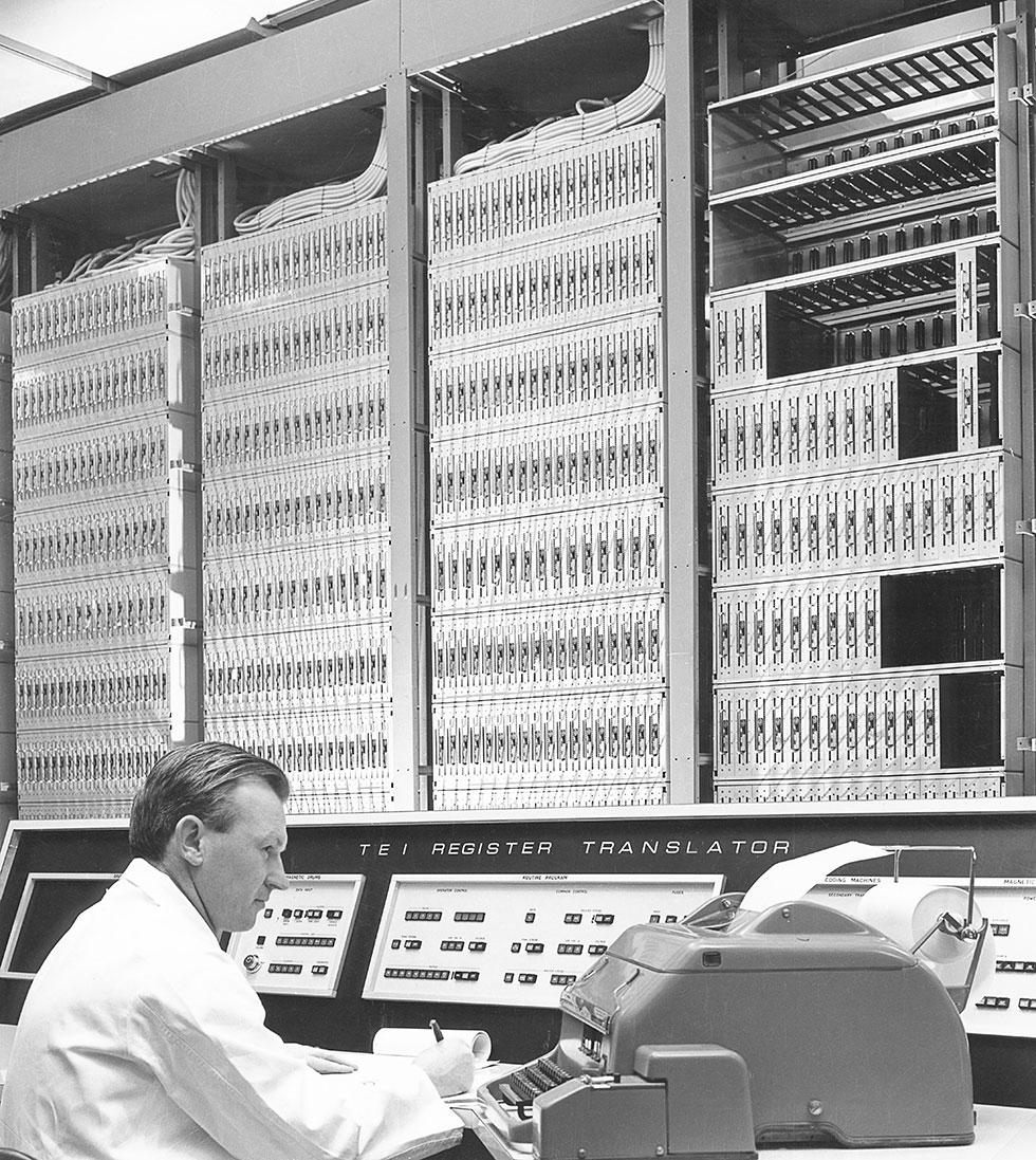 A man in a white shop-coat sits at a desk beside a large printer and console with switches.