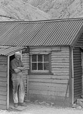 A man standing in the doorway of a timber hut. Denuded hills behind.