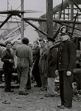 Men dressed in overalls and a policeman standing within the ruins of a fire damaged warehouse.