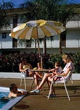 A man leaning against the side of a swimming. Two women with children seated under an umbrella.