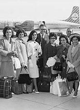 6 women and 2 men stand before a railing with a jet passenger aircraft behind them.