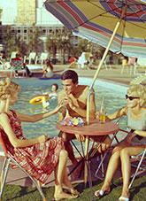 Two women and a man seated at a table under a beach umbrella beside a pool.