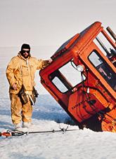 Man in yellow leaning against orange grader wedged in ice crevice.