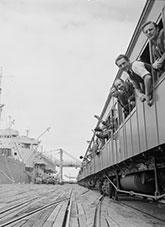 People leaning from rail carriage windows. Bridge and ship in the background.