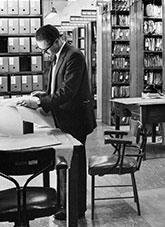 An archives officer examining plans. Files on compactus shelves behind him.