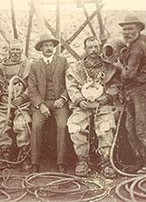 Group photo of 5 men. 2 men wearing diving suits and 2 men holding their diving helmets. A fifth man wearing a suit.