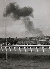 View across a grass racecourse toward smoke rising above buildings.