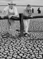 2 farmers leaning against a timber rail fence examining the parched ground of a dried-up dam.