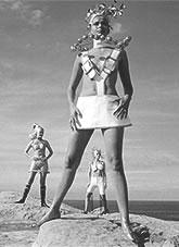 Three women standing on a sandstone cliff above the ocean dressed in futuristic fashion.