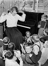A class of young children gathered around a teacher seated at a piano shaking her hands.
