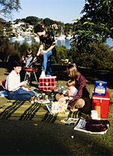 3 people and 2 small chidlren with a BBQ and picnic blanket on lawn with boats in the background.