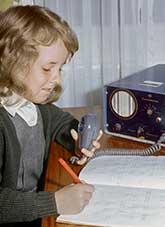 A young student writing in her study book while holding the microphone attached to a small radio tranceiver.