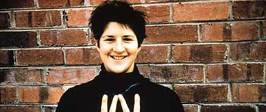 Dawn Fraser wearing a black long sleeved top standing before a brick wall making the V for victory sign with both hands.