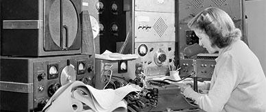 A woman talks into a microphone at a desk supporting a stack of radio equipment.