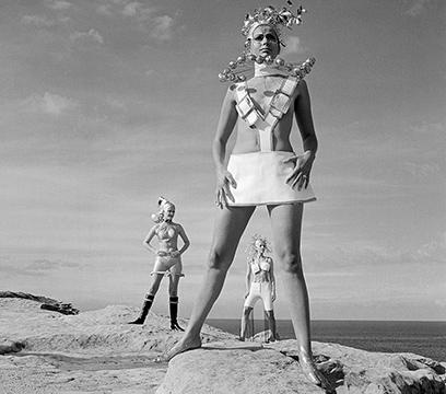 Three women standing on a sandstone cliff above the ocean dressed in futuristic fashion.