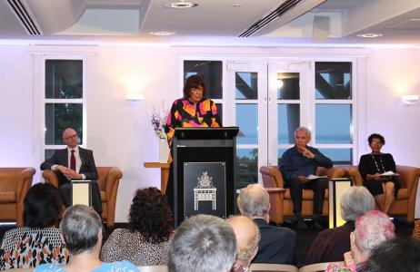 Audience listening to a woman speaking from behind a lectern and participants seated beside her.