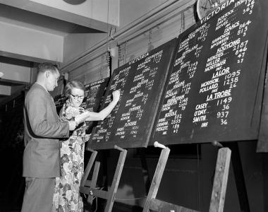 A man and a woman update tally boards mounted on easels using chalk. 