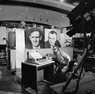 Journalists on a raised platform sit behind large photographs of Gough Whitlam and John Gorton. A man wearing headphones watches a print-out from a Telex machine.