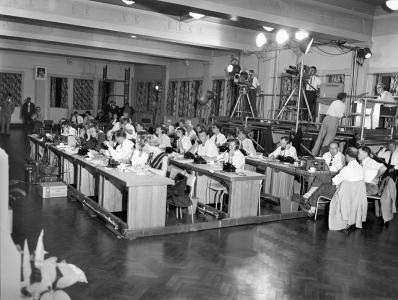 Television cameras on tripods and numerous people seated at desks.