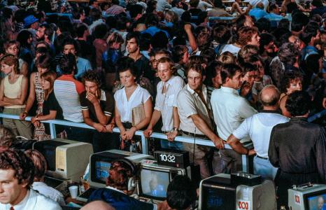 People leaning on a railing behind TV monitors.