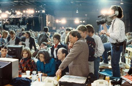 People crowded around a monitor while a video camera operator stands on a chair recording.