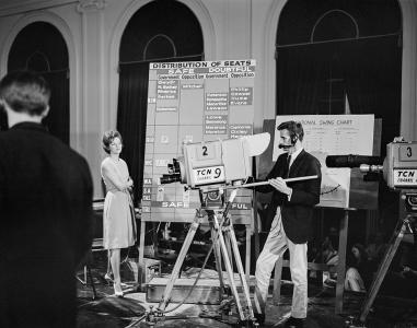 A female newsreader and male camera operator beside a tally board.