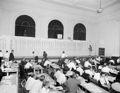 People seated at desks. A tally board beneath large arched windows at the side of the room.