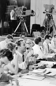 A row of people with communication headsets. Large television cameras in the background.
